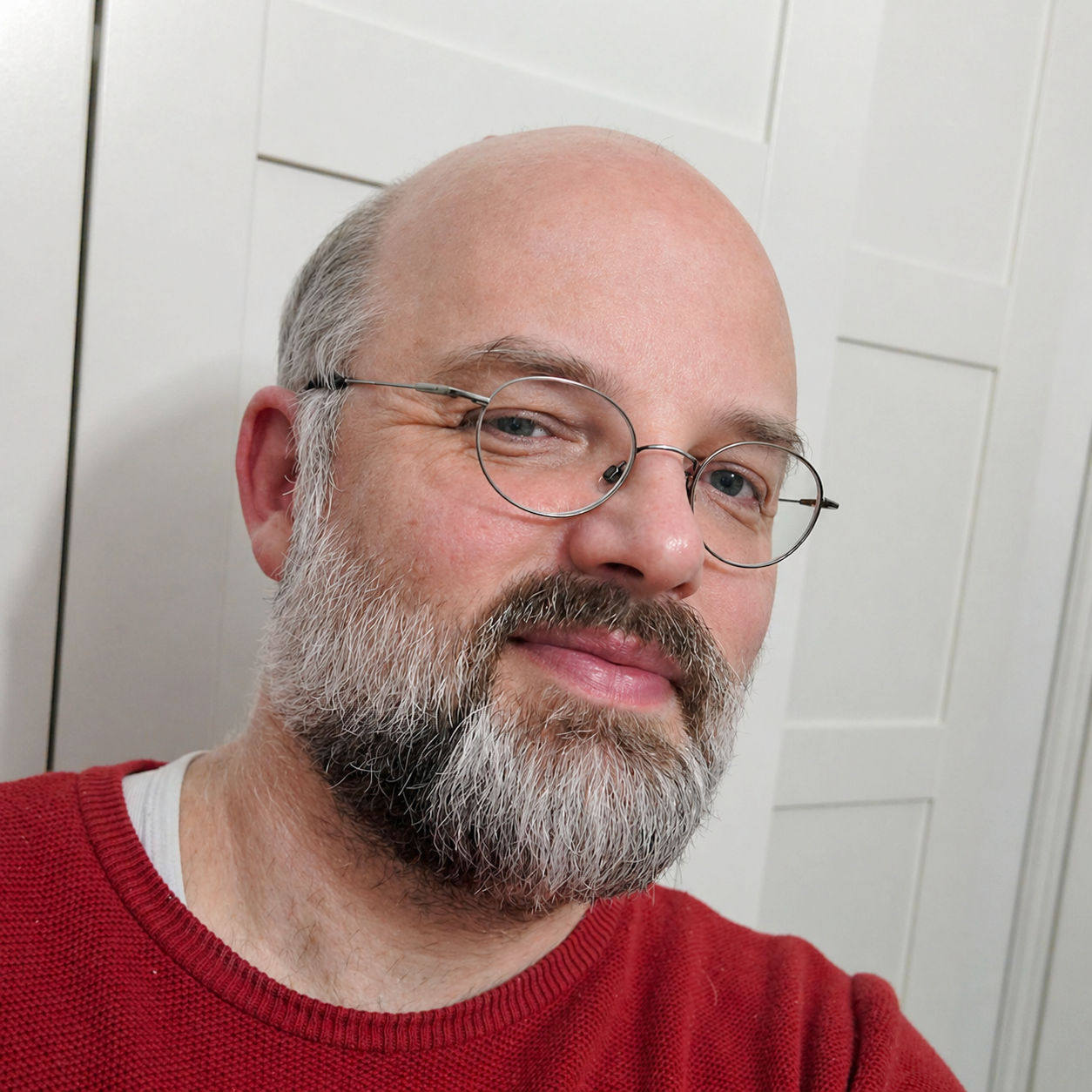 Alexander Rüther mit Brille, Bart und rotem Pullover vor hellem Hintergrund.